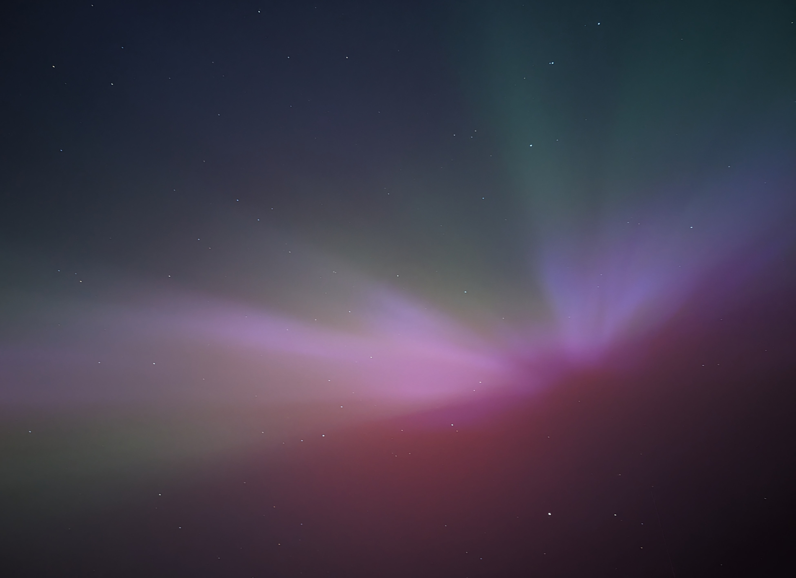 A photo of the Aurora Borealis visible in the night sky. A horizontal glowing pink strip with cascading light beams of purple and green. The stars are visible behind it.