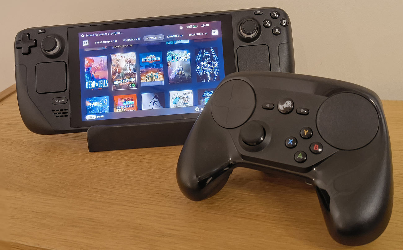 On top of a wooden surface, a Steam Deck is propped up on a stand, its screen is turned on and is showing a selection of games ready to play. To the right of it is an original Steam Controller.