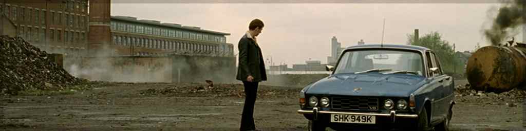 A cropped screenshot from 'Life on Mars' episode one. In the centre, a man in a leather jacket gazes down at a blue car from the 70s. He stands in the middle of an industrial estate, grey skies and an old brick factory behind him.