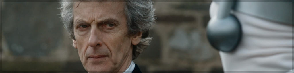 A cropped screenshot from an episode of 'Doctor Who'. The Twelfth Doctor played by Peter Capaldi, glares to a humanoid creature, a 'Cyberman' blurred in the foreground – You can't see its face. The Cyberman's head is covered in a white fabric, with metal tubing extruding from the side of its head. They stand in front of a building with a large stone wall.